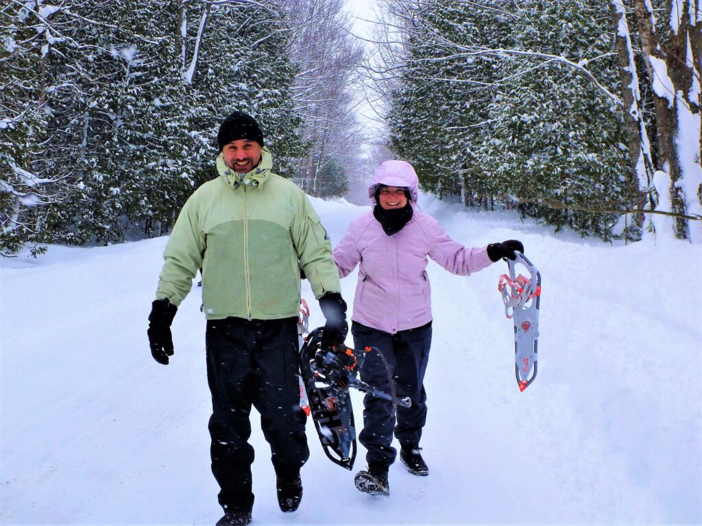 Couple walking a snow-covered trail while holding snowshoes.